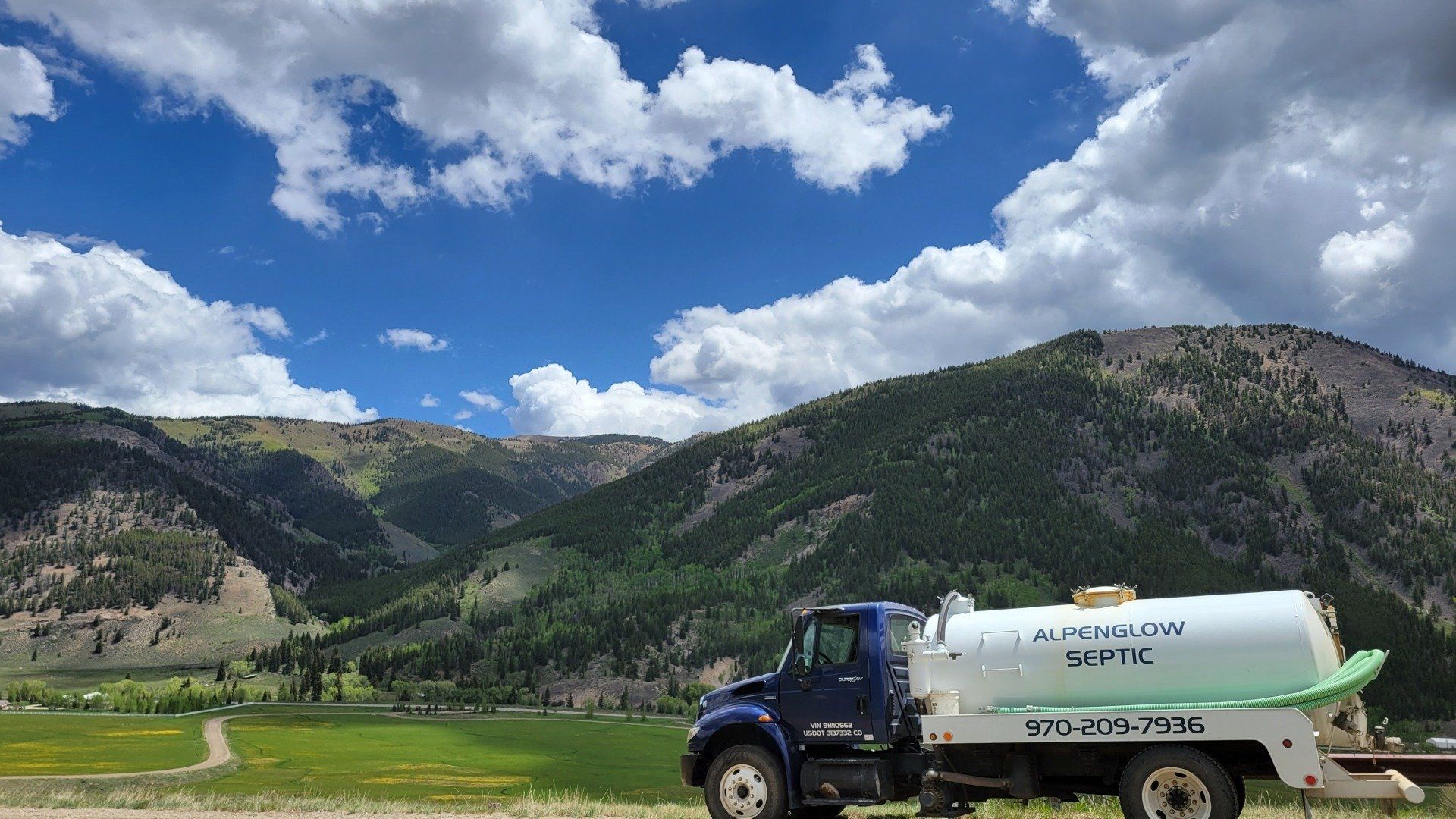 Blue tanker truck spraying fields in a green valley beneath rocky mountains and a cloudy sky