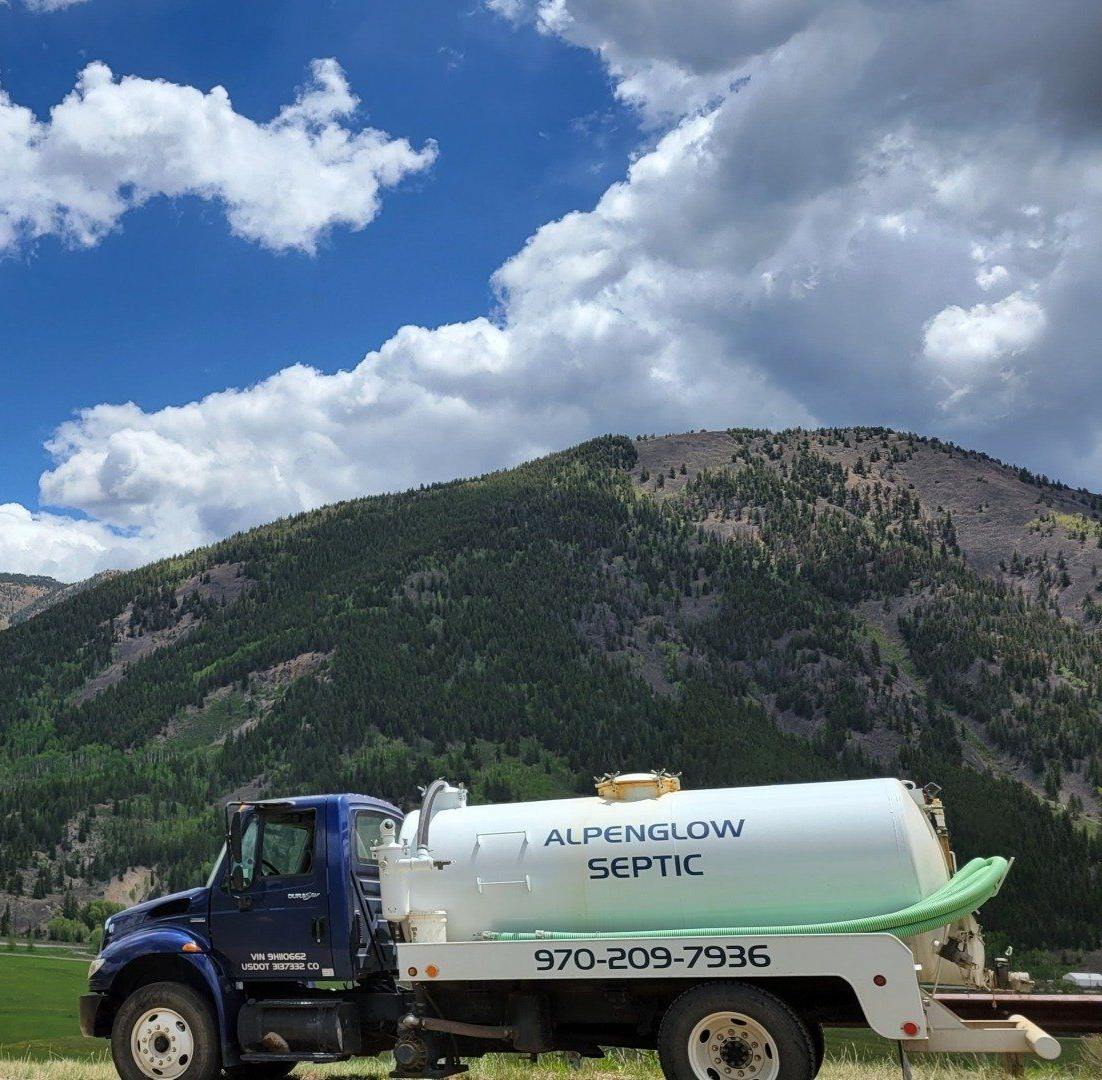 Blue septic truck parked in front of a forested mountain under a cloudy sky