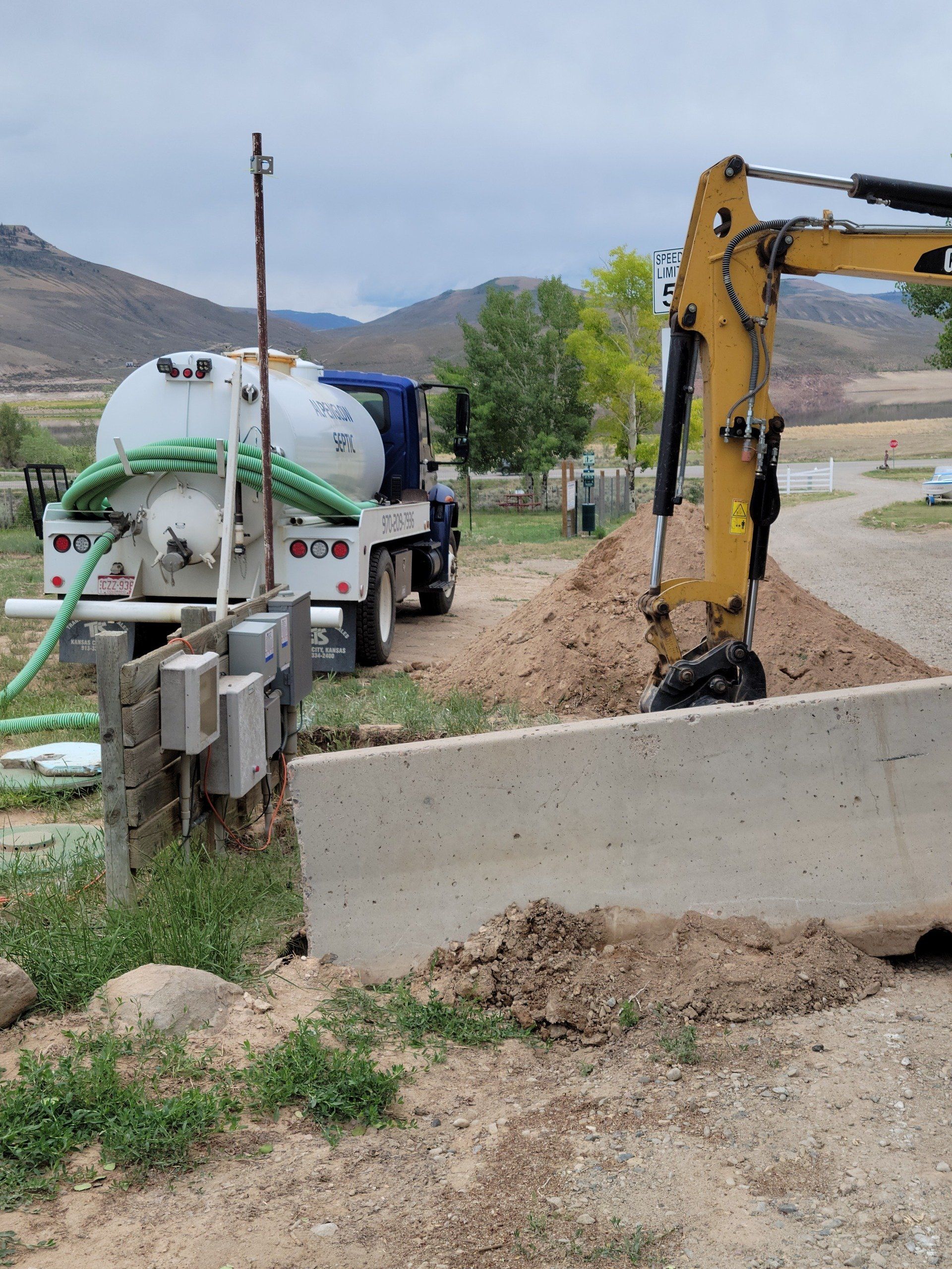 Excavator digging beside a concrete barrier with a tanker truck on a rural roadside.