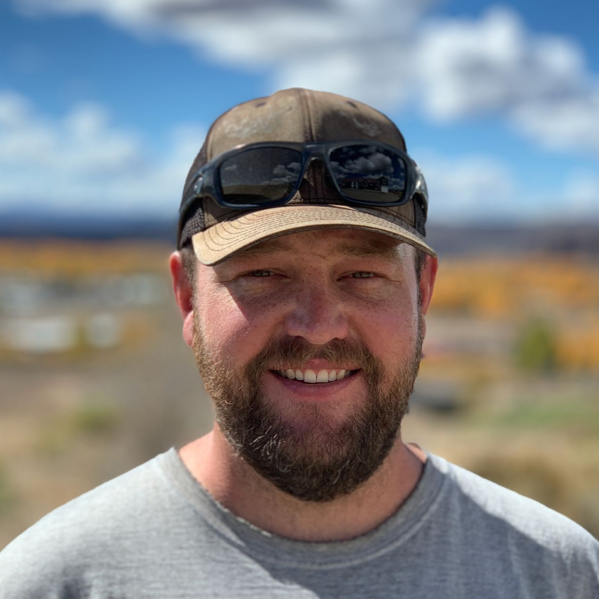Smiling bearded man in sunglasses and cap outdoors with blurred mountains and blue sky background