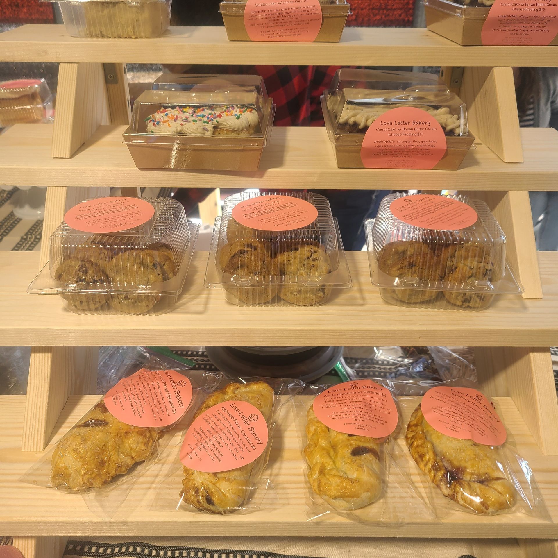 Stacked trays of packaged pastries with pink labels on a wooden display shelf.