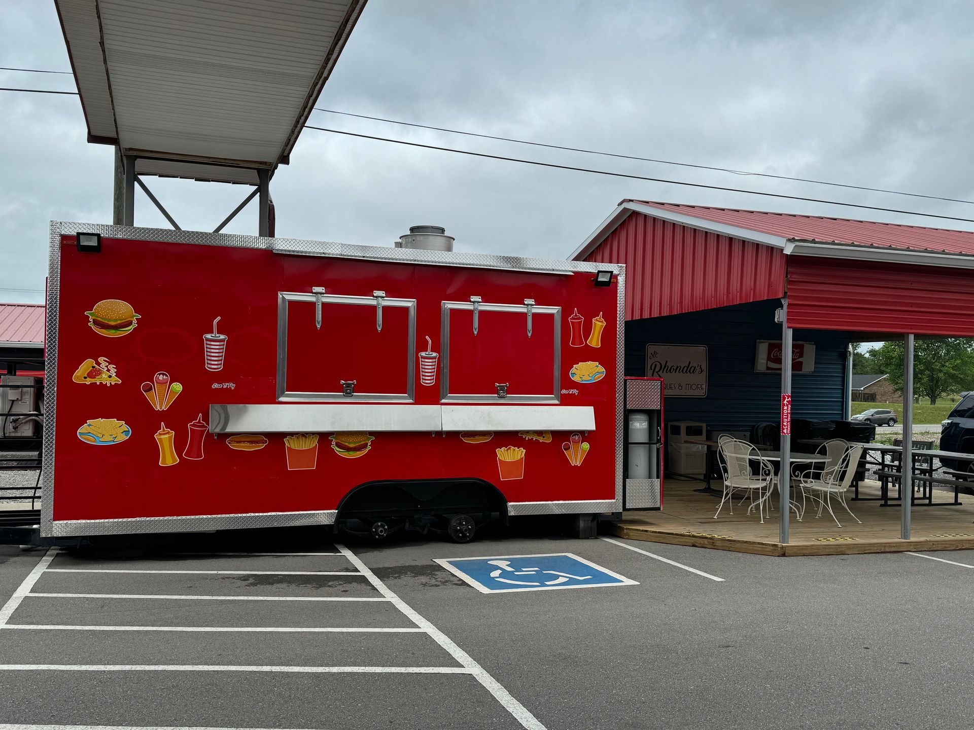 Red food truck parked in a lot beside a red building, with serving windows and a blue accessible space nearby.
