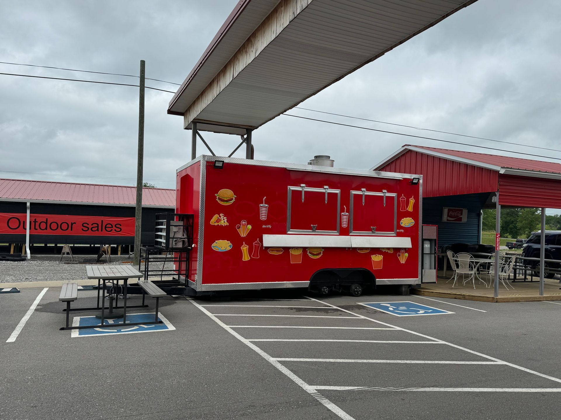 Red food trailer at a covered outdoor market with picnic tables and parking spaces