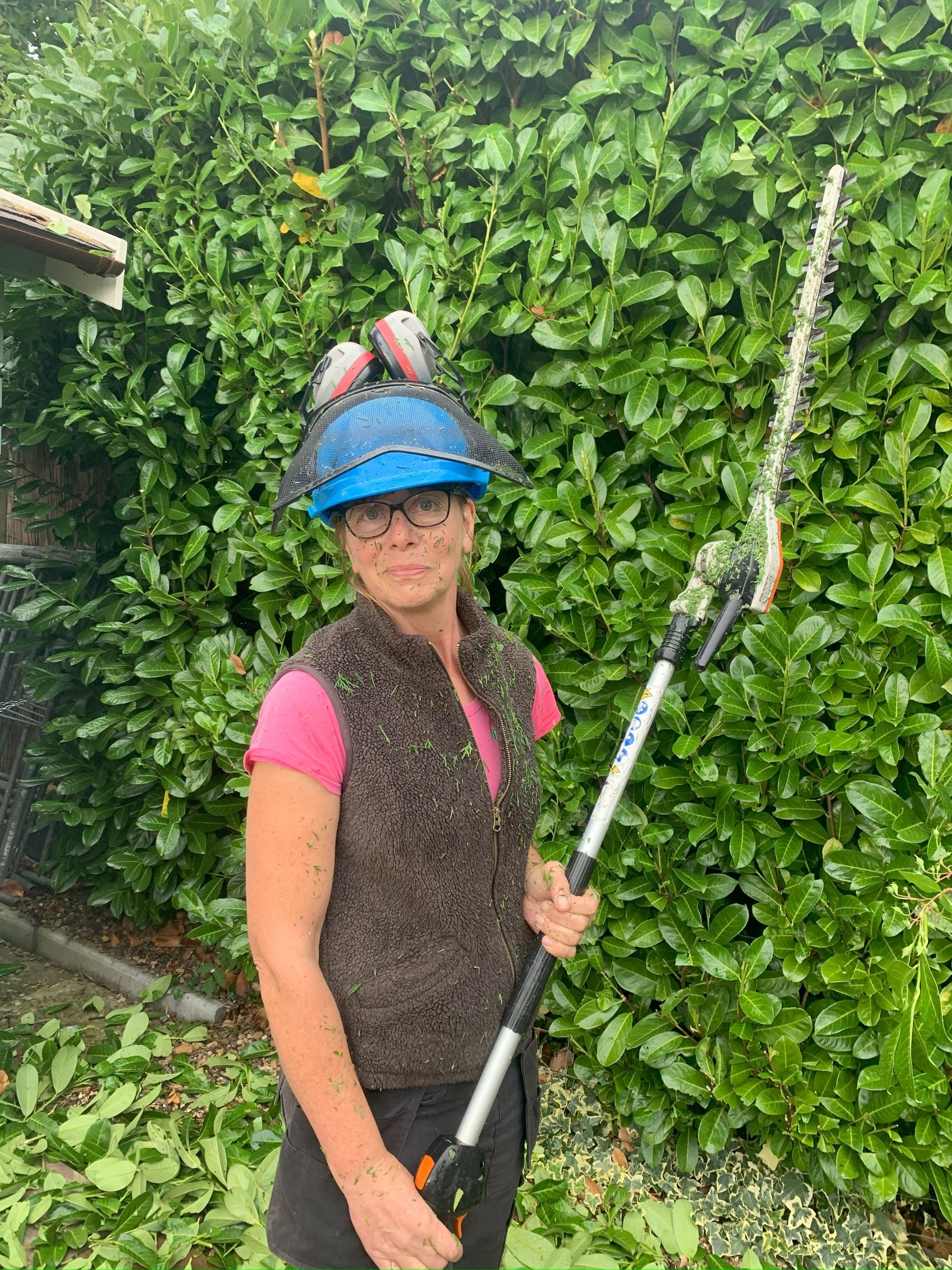 A woman is standing in front of a hedge holding a hedge trimmer.