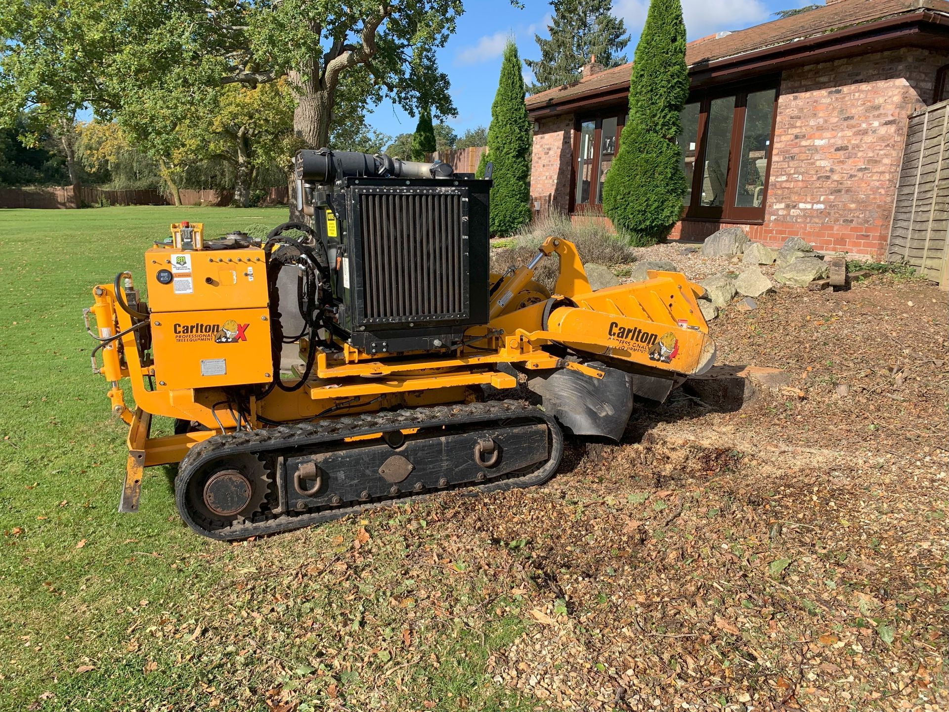 A yellow stump grinder is sitting in front of a brick house.