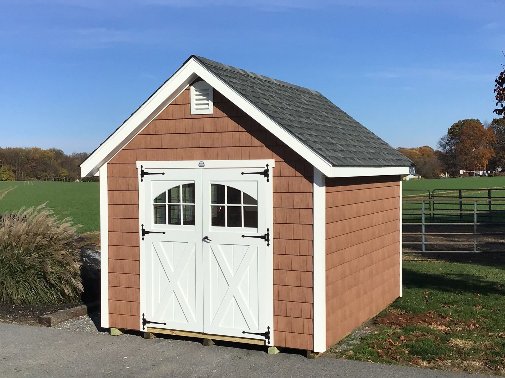 Classic Garden Cottage Shed with light brown wood siding, white double doors with windows, white trim, a white vent, roof overhangs on all sides, and a dark gray asphalt roof.