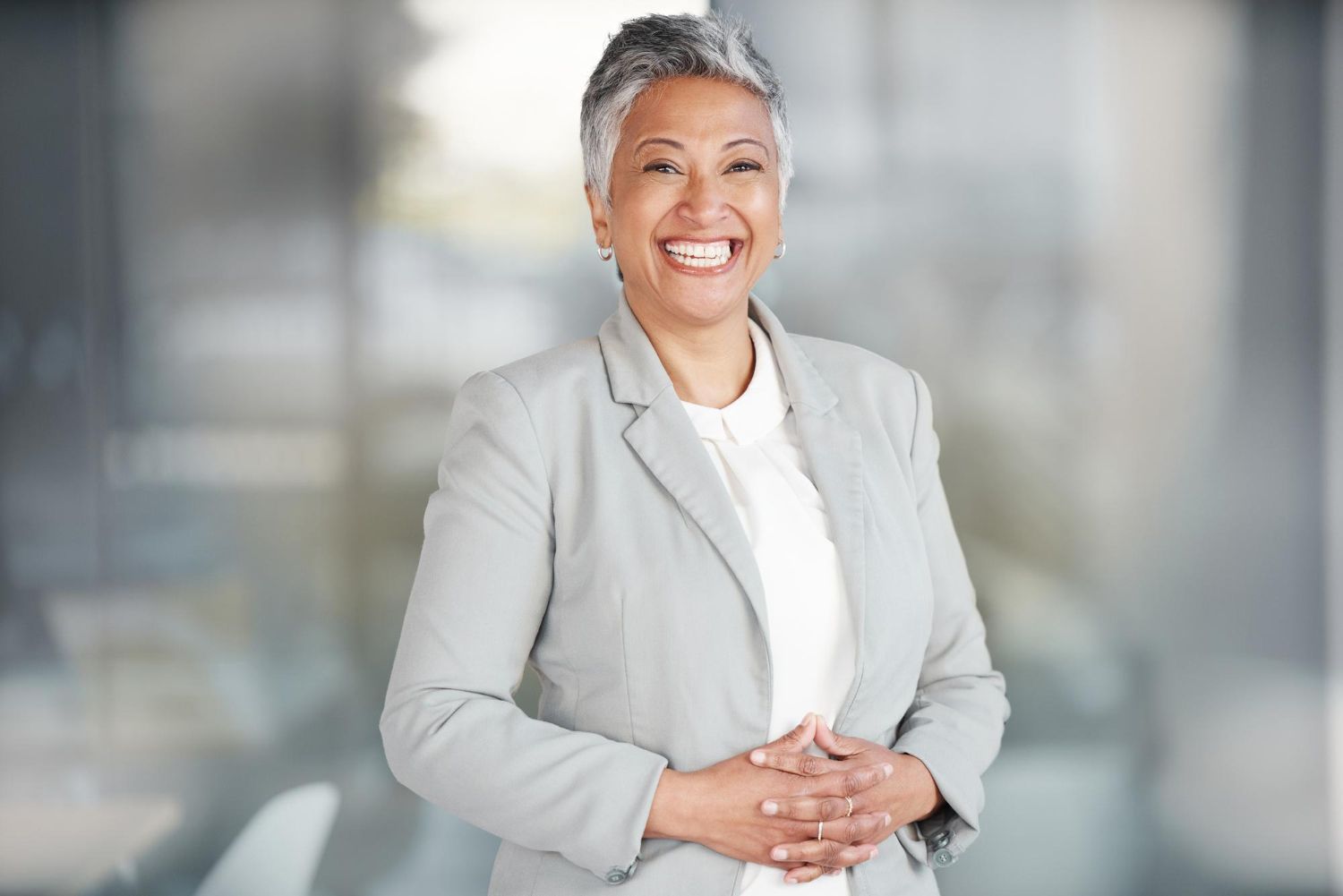 Smiling person in a light gray blazer standing in a bright office with hands clasped.