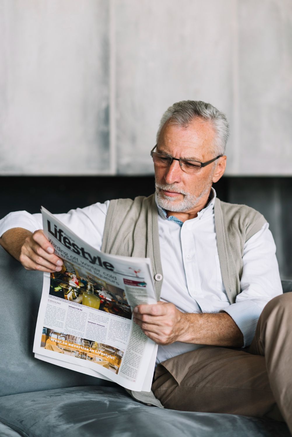Man reading a newspaper on a couch, wearing glasses and a light vest, in a modern indoor setting
