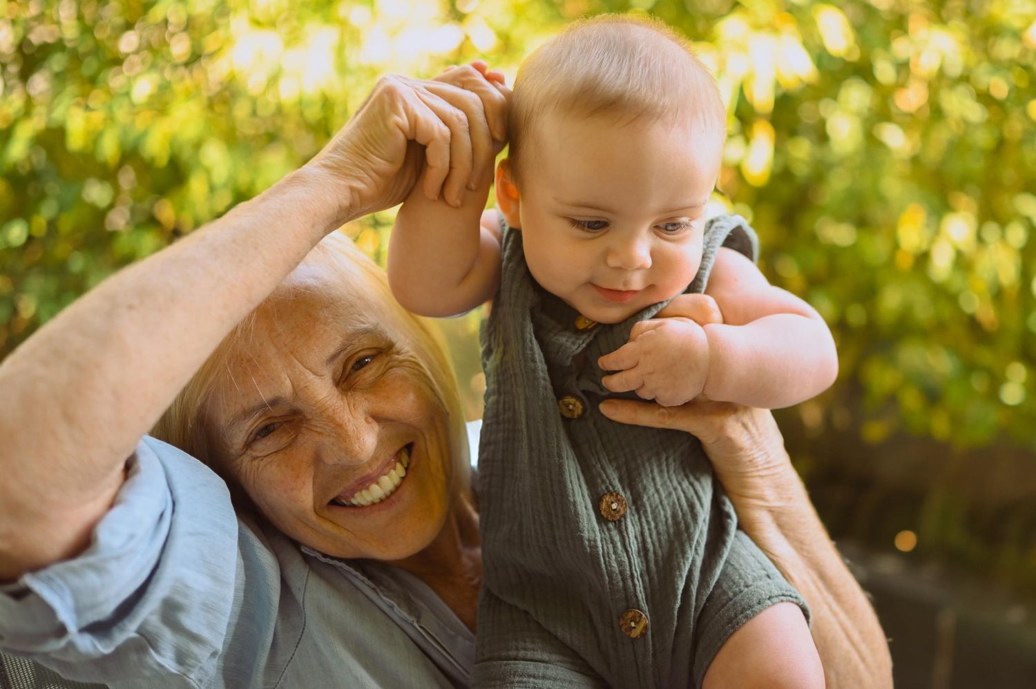 Older adult smiling while holding a baby outdoors in warm sunlight