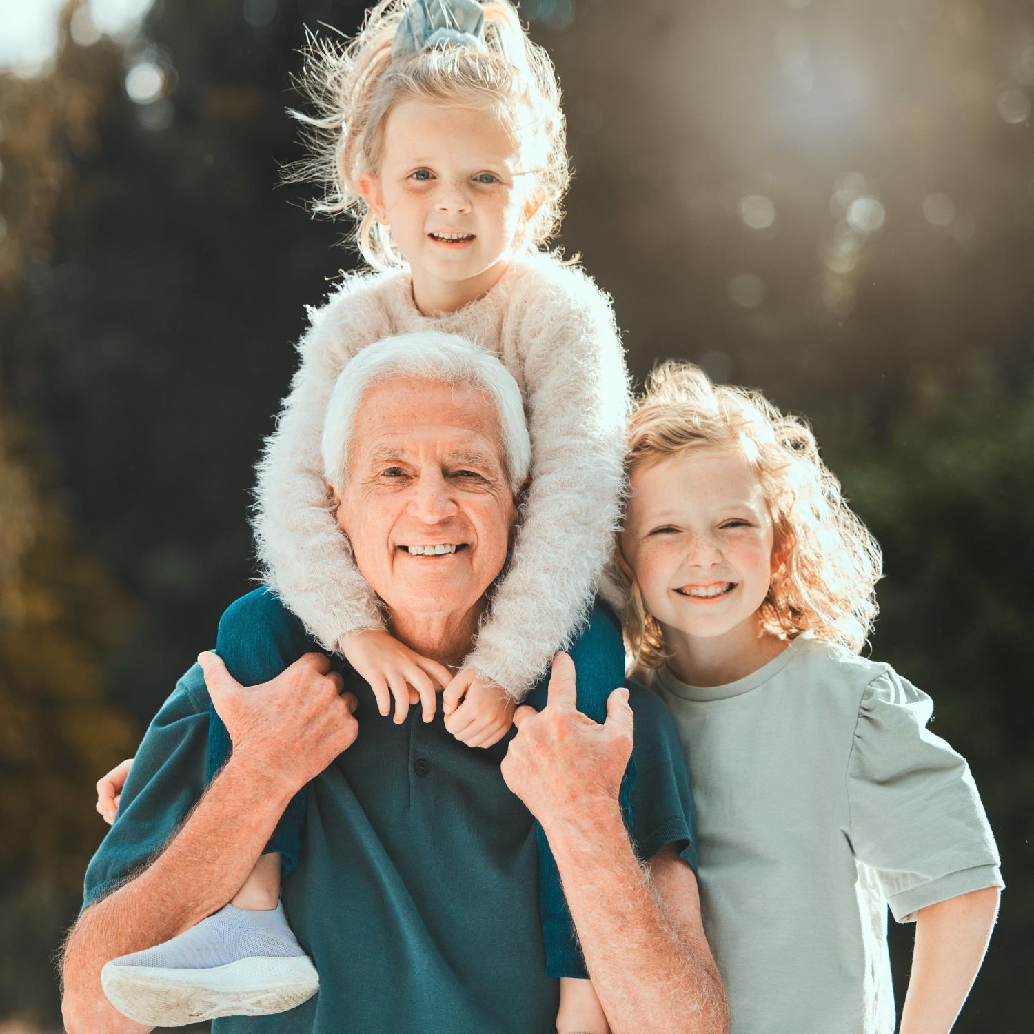 Smiling elderly man carrying two children on his shoulders outdoors in sunlight