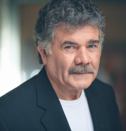 Portrait of a man with gray hair and mustache, wearing a dark blazer and white shirt, looking at the camera.