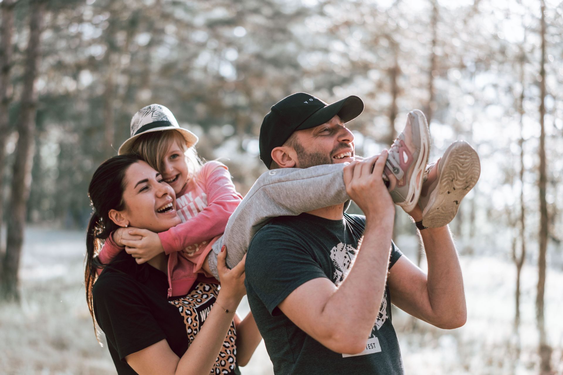 Two adults carrying a smiling child on their shoulders in a wooded outdoor setting