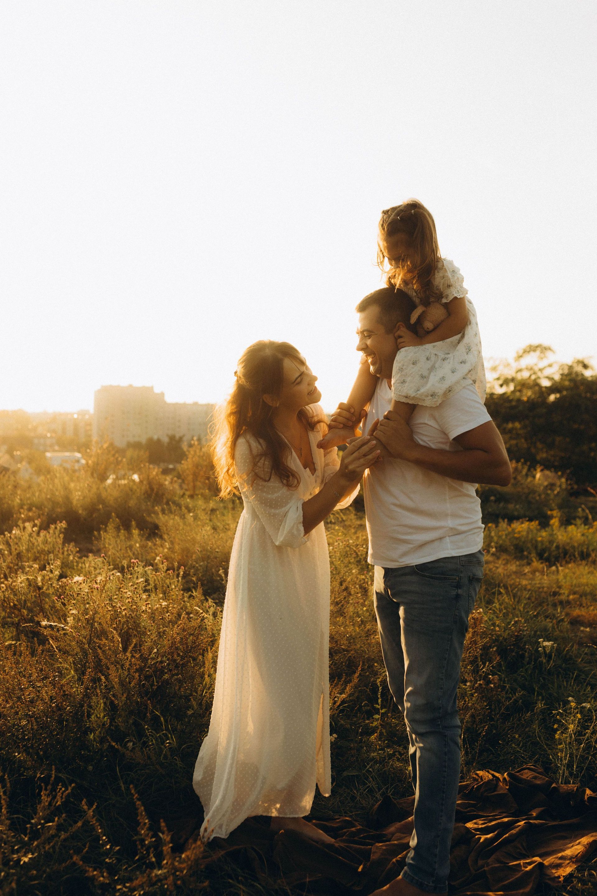 Family of three in a sunlit field at sunset, with a child sitting on an adult's shoulders and another adult holding hands nearby