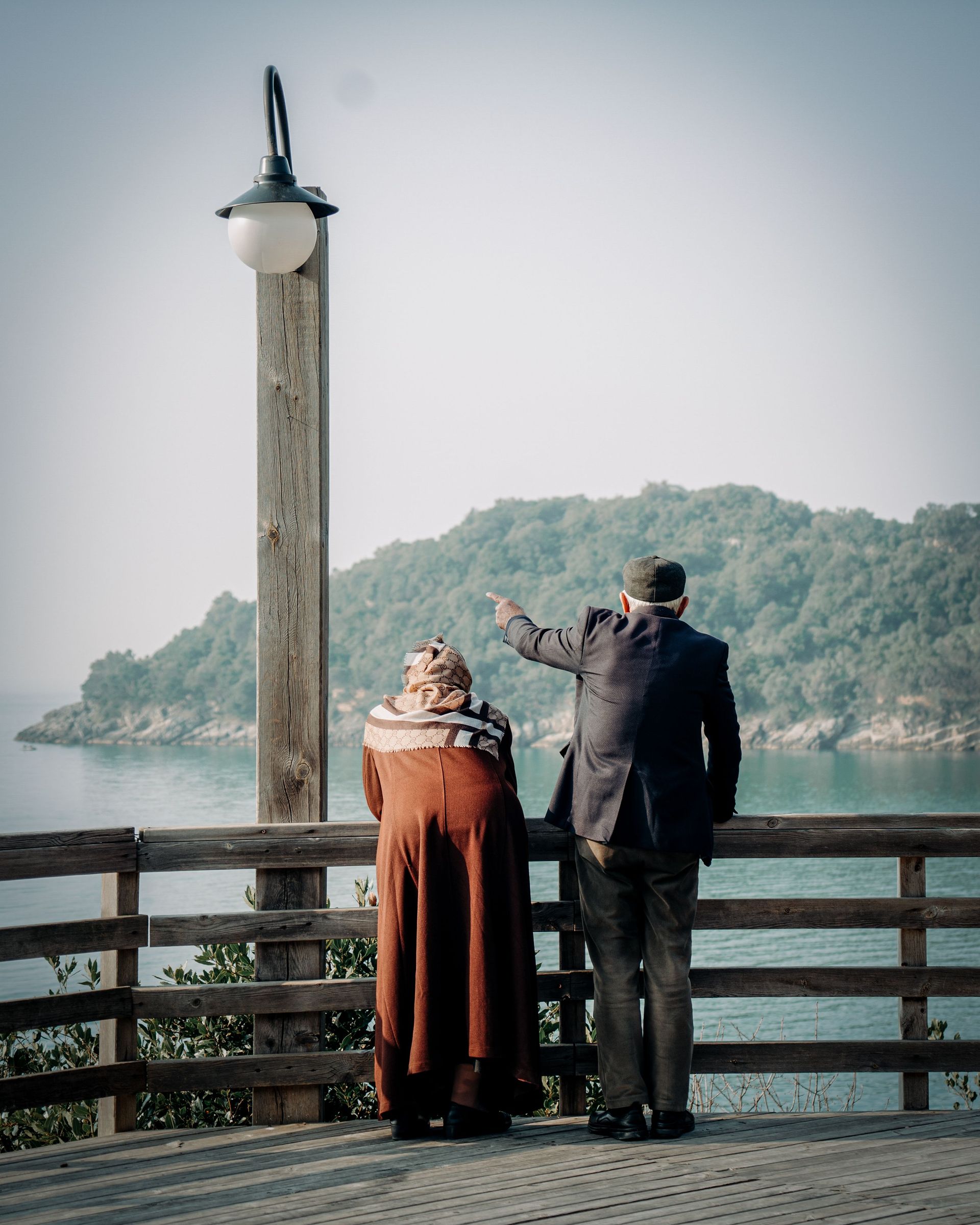 Two people stand on a seaside deck, looking at a green island across calm water.