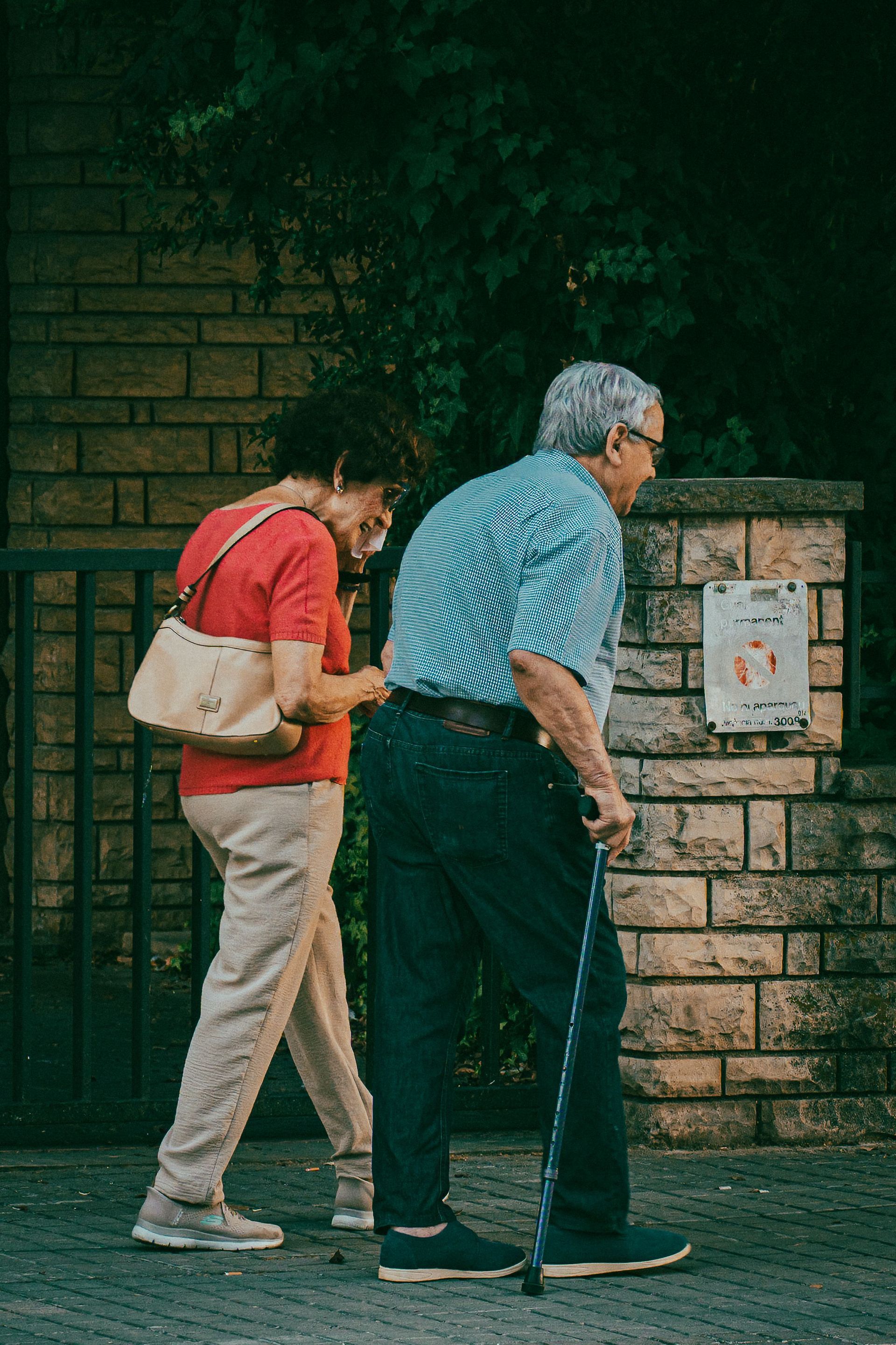 Two people walking past a brick wall and notice board, one in a red shirt with a beige bag.