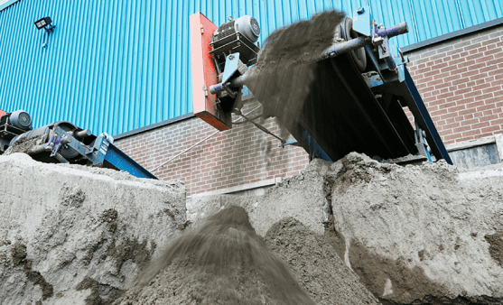 Conveyor belt depositing sand onto a large pile against a blue and brick industrial building.