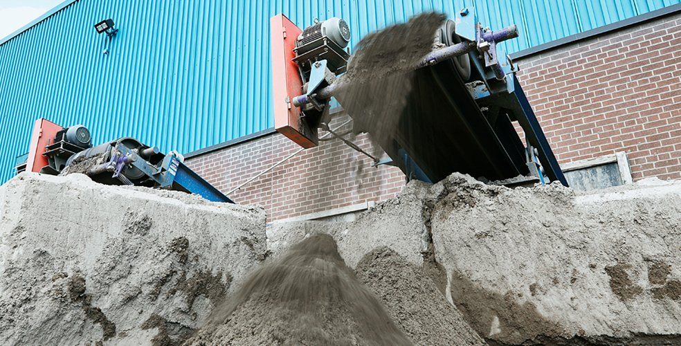 Industrial machine dumping sand into a pile between concrete blocks. Blue and red machinery, brick building backdrop.
