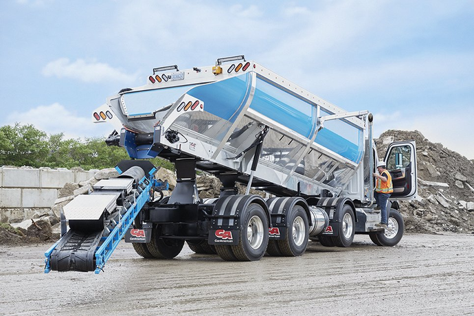A dump truck with a blue and silver bed is dispensing material onto a conveyor belt. A person is standing in the truck's cab.