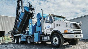 White and blue vacuum truck with raised tank, parked outside a building.