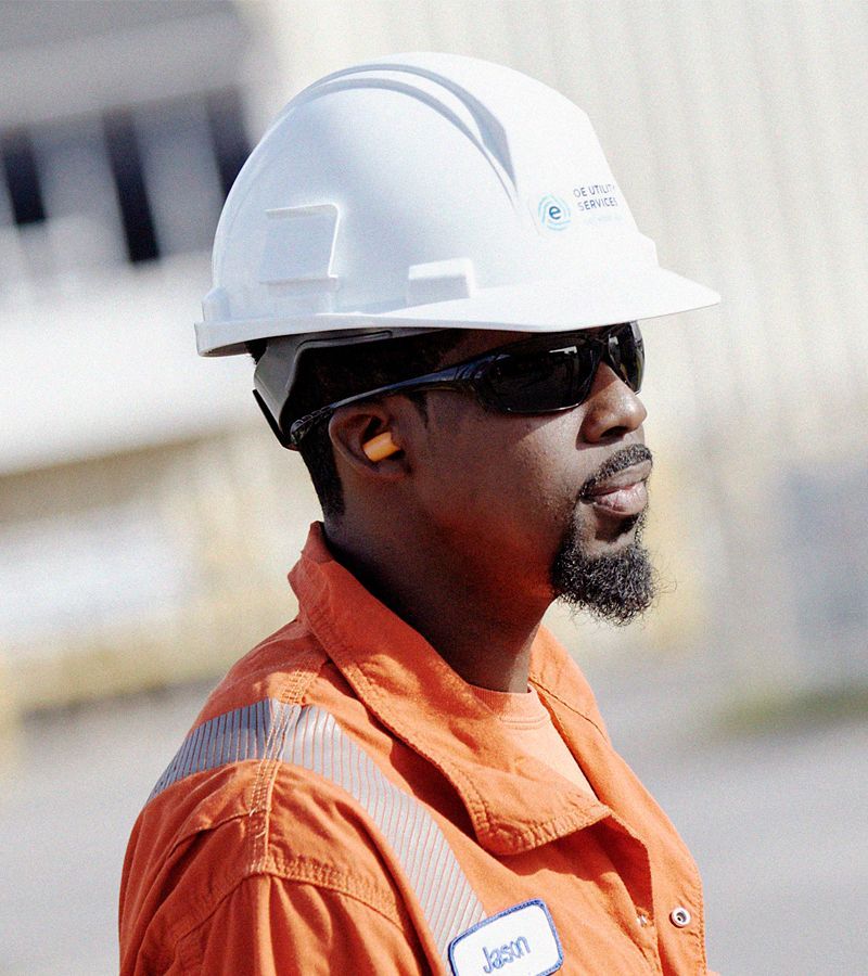 Person in orange workwear, safety glasses, hard hat, and earplugs outdoors, looking right.