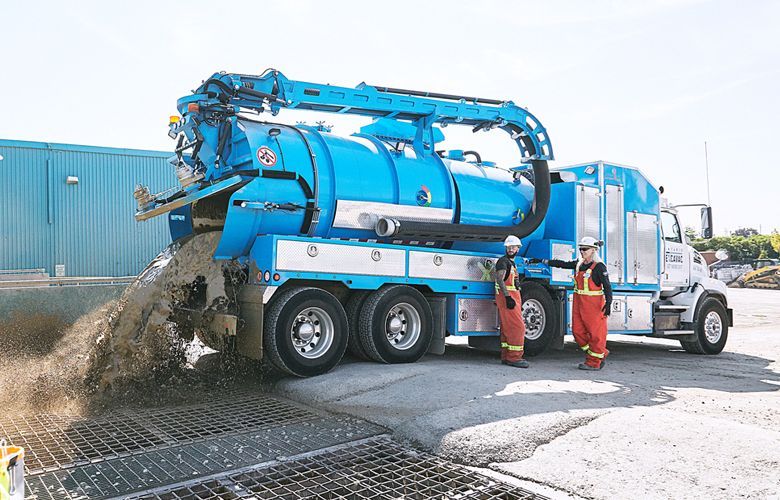 Two workers stand next to a large blue vacuum truck, discharging debris onto a grate.