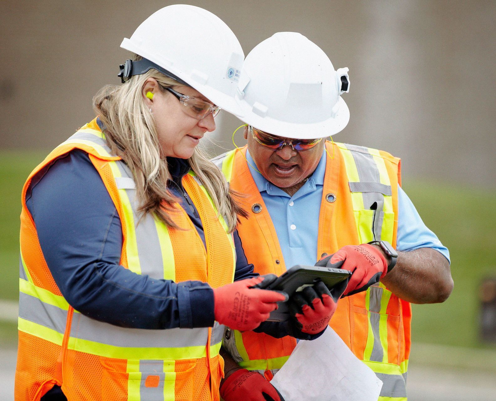 Two people in hard hats and safety vests looking at a tablet outdoors.