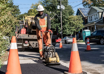 Construction worker compacting asphalt with a plate compactor on a residential street. Orange safety cones.