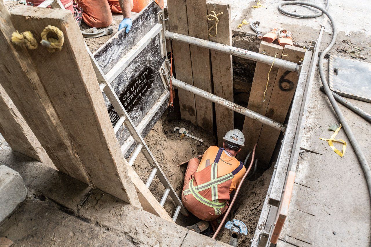 Construction worker in trench, wearing safety gear. Wooden shoring supports the walls, ladder inside.