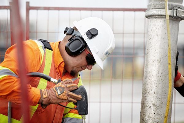 Construction worker in safety gear using a tool near a metal pipe.