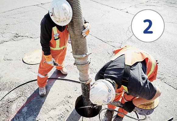 Two workers in orange vests and hard hats, one holding a hose, inspecting a manhole.