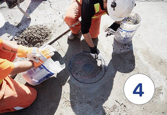 Workers in orange vests pour concrete mix around a manhole cover on a concrete surface.