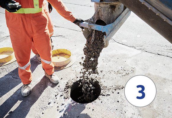 Person in orange jumpsuit pouring concrete into a hole in pavement.