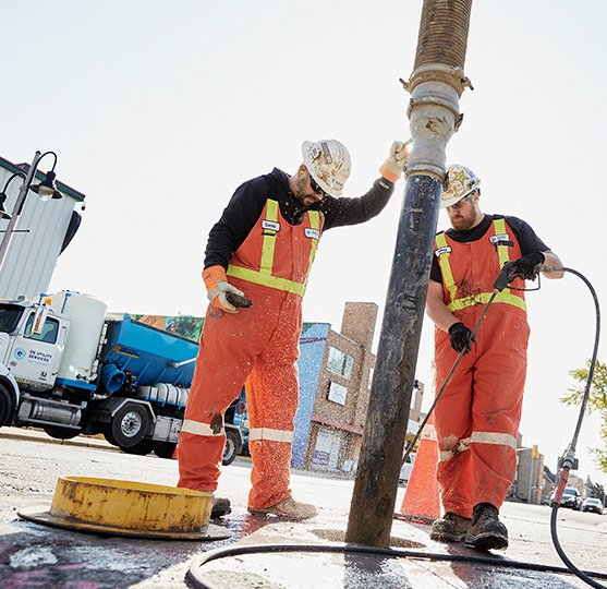 Two workers in orange coveralls near a utility pole, one holding a hose. A truck is visible in the background.