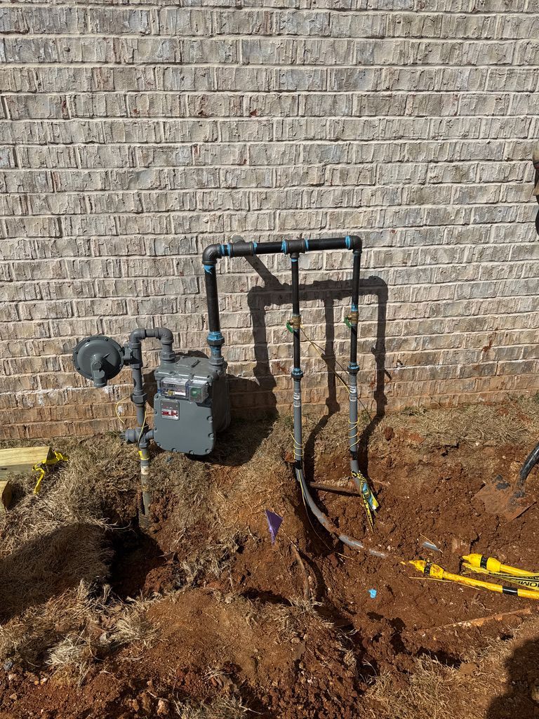Outdoor gas meter and pipes beside a brick wall, with dirt ground and a trench in front.