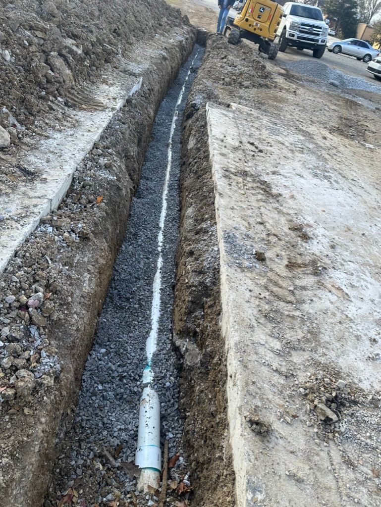 Trenched utility pipe laid in a muddy roadside ditch beside snow, with construction vehicles in the background