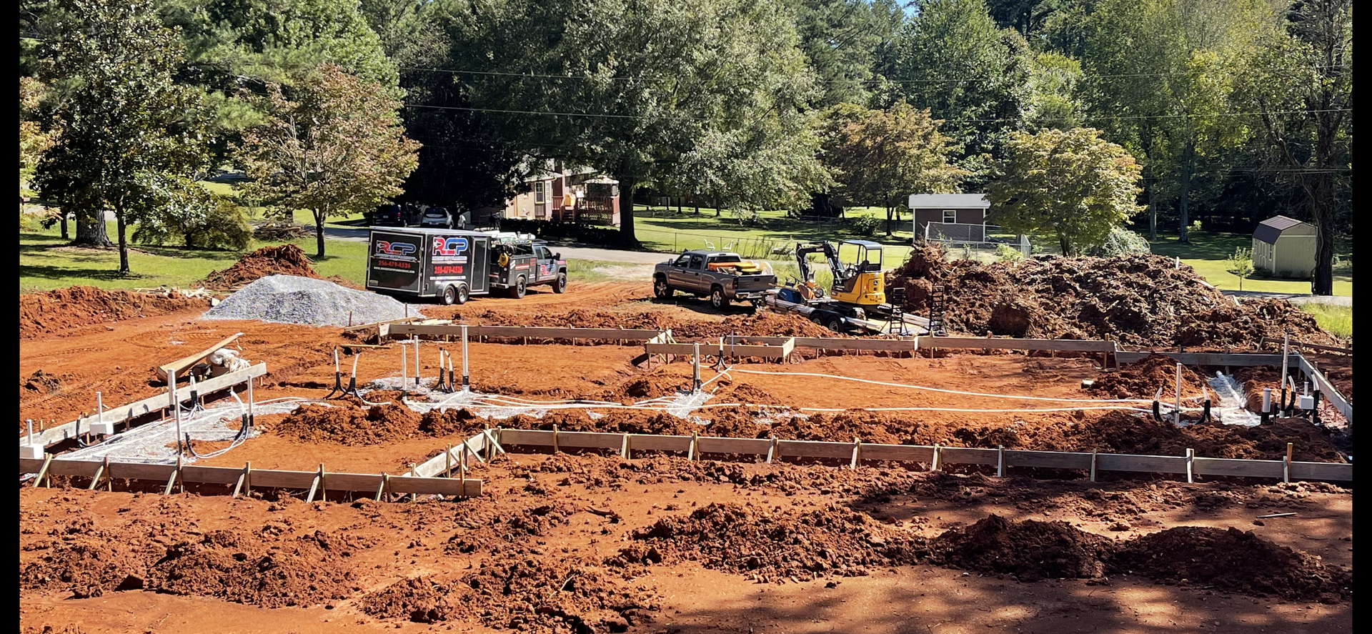 Construction site with orange soil, concrete forms, and piles of debris beside trees and parked vehicles