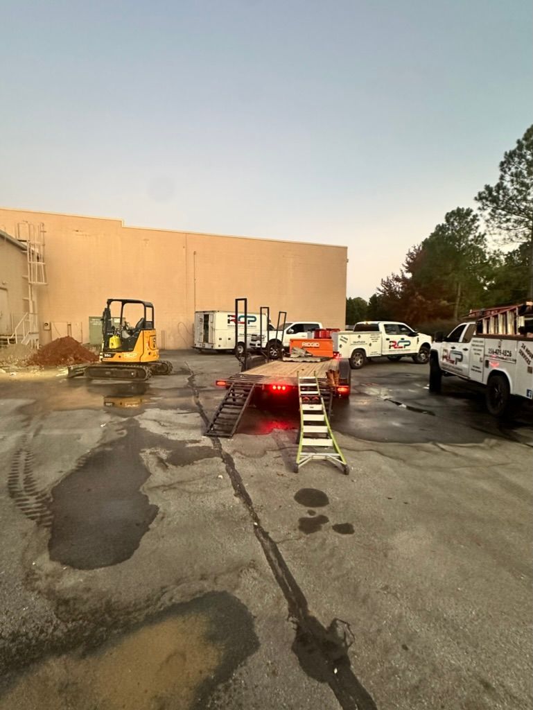 Construction equipment and work trucks on a cracked asphalt lot beside a beige building at dusk