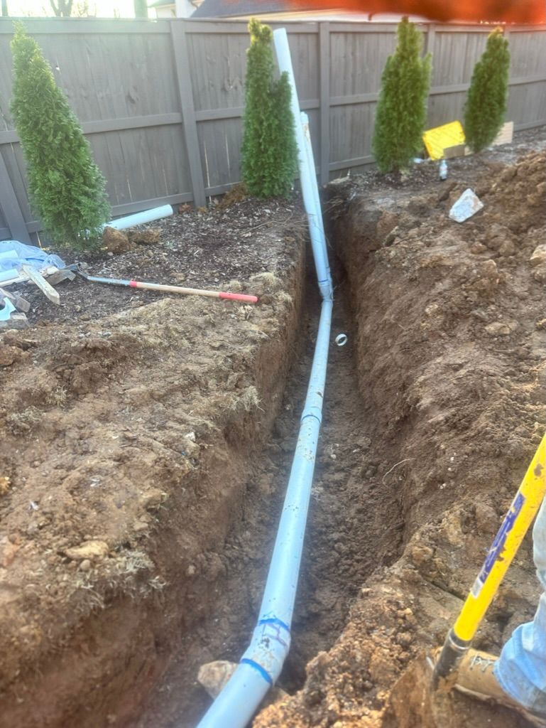 Trench with a blue pipe installed in a backyard beside a wooden fence and shrubs