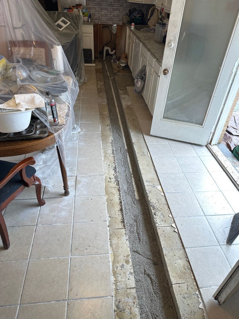 Narrow indoor hallway with beige tile floor, exposed trench, and doorway leading to a kitchen area