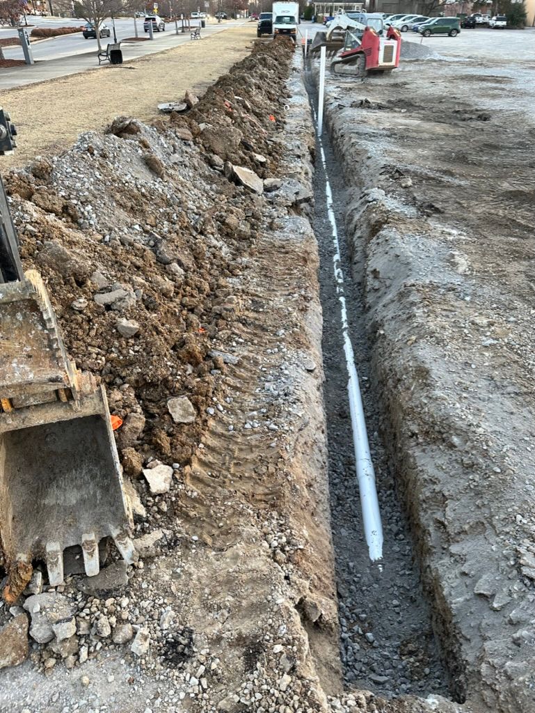 Trench excavation beside a road, with a white pipe laid in a narrow ditch and an excavator bucket nearby.