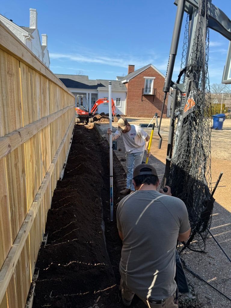 Workers digging a trench beside a wooden fence on a sunny construction site