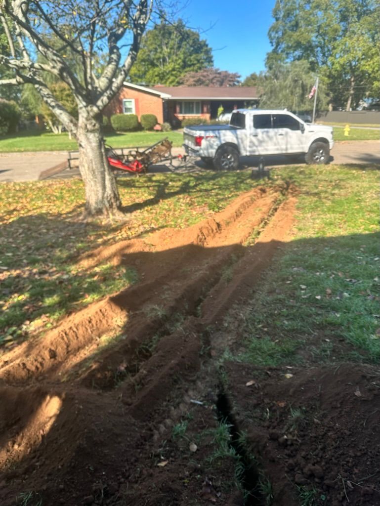 Dirt driveway with deep tire ruts beside a white pickup truck in a yard with a brick house and trees.