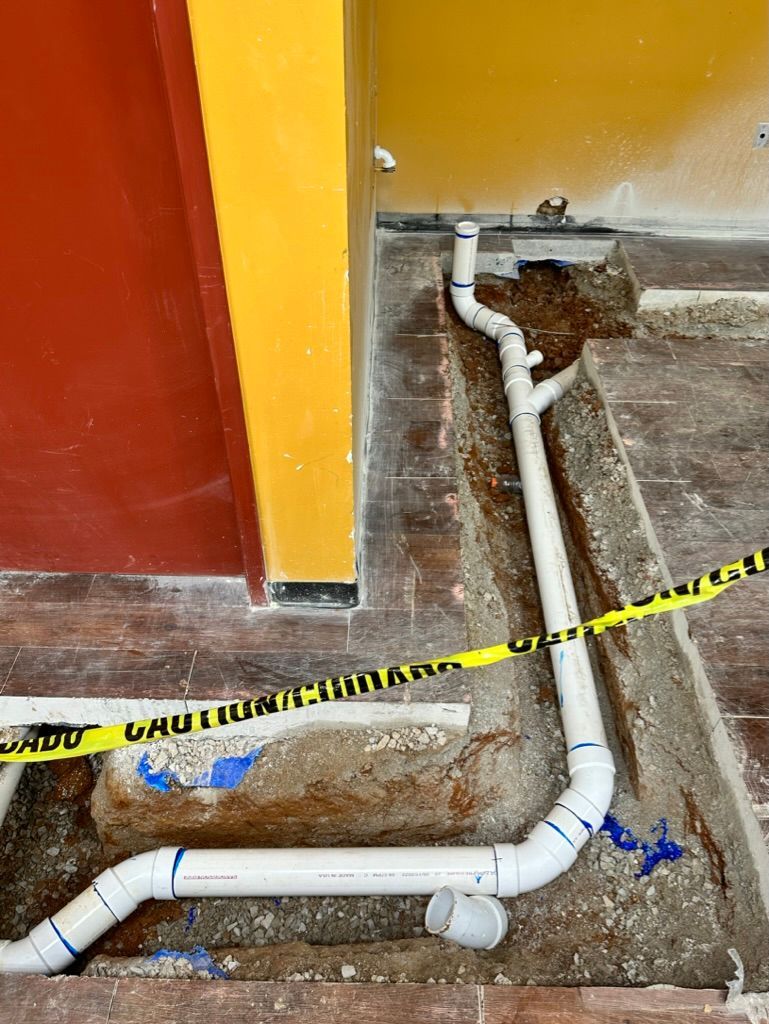 Exposed white PVC plumbing pipes in a damaged floor area beside a red and yellow wall, marked with caution tape