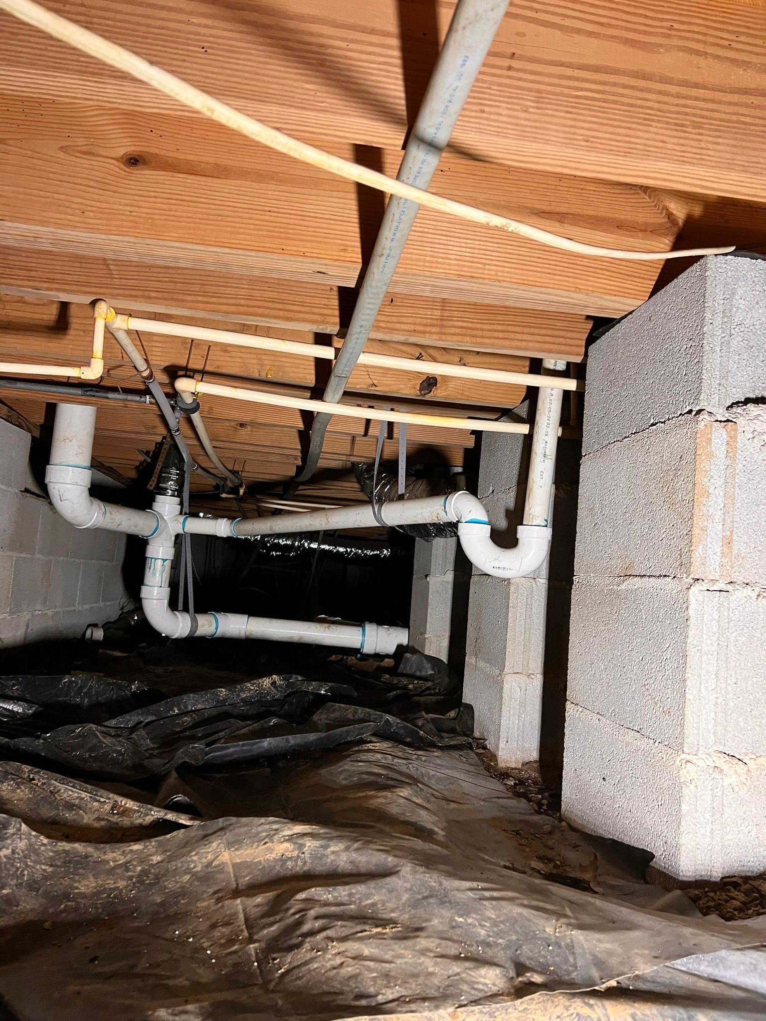 Crawlspace under a house with exposed pipes, insulation, and a plastic-covered dirt floor