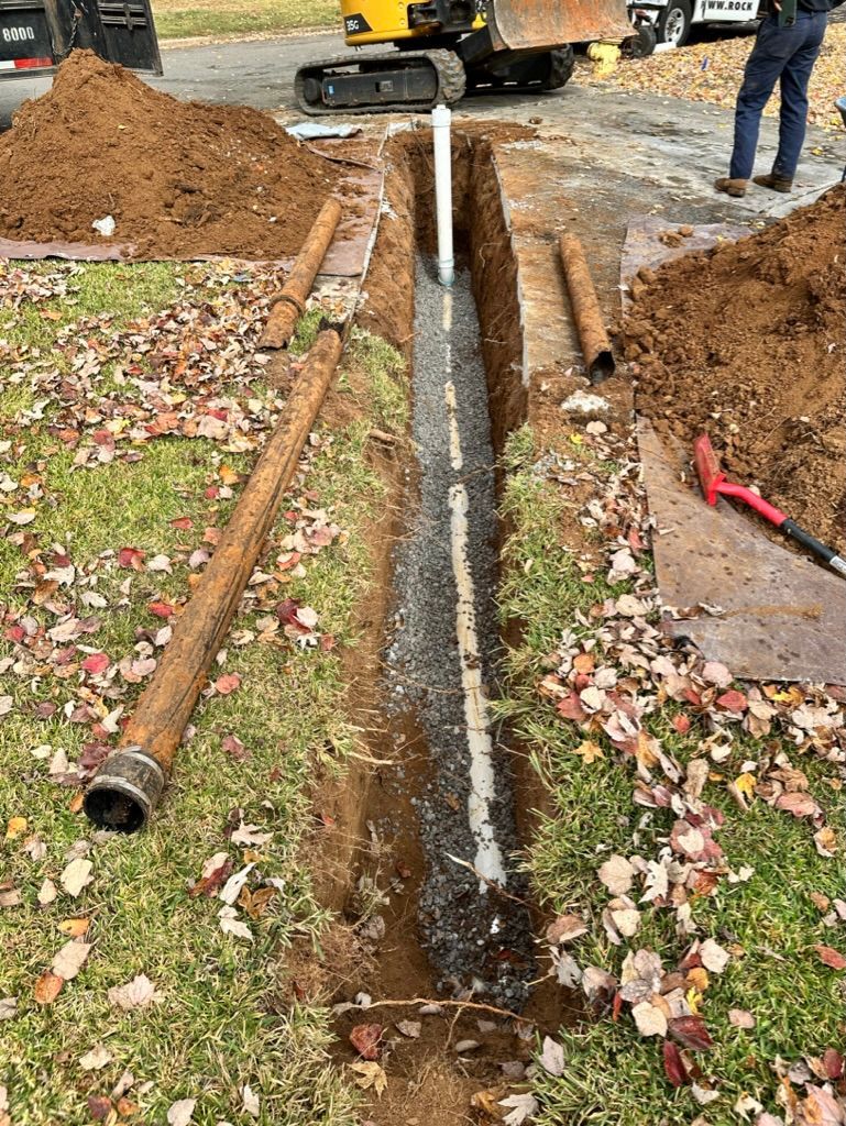 Open trench with exposed pipe and muddy dirt piles beside a sidewalk during utility work