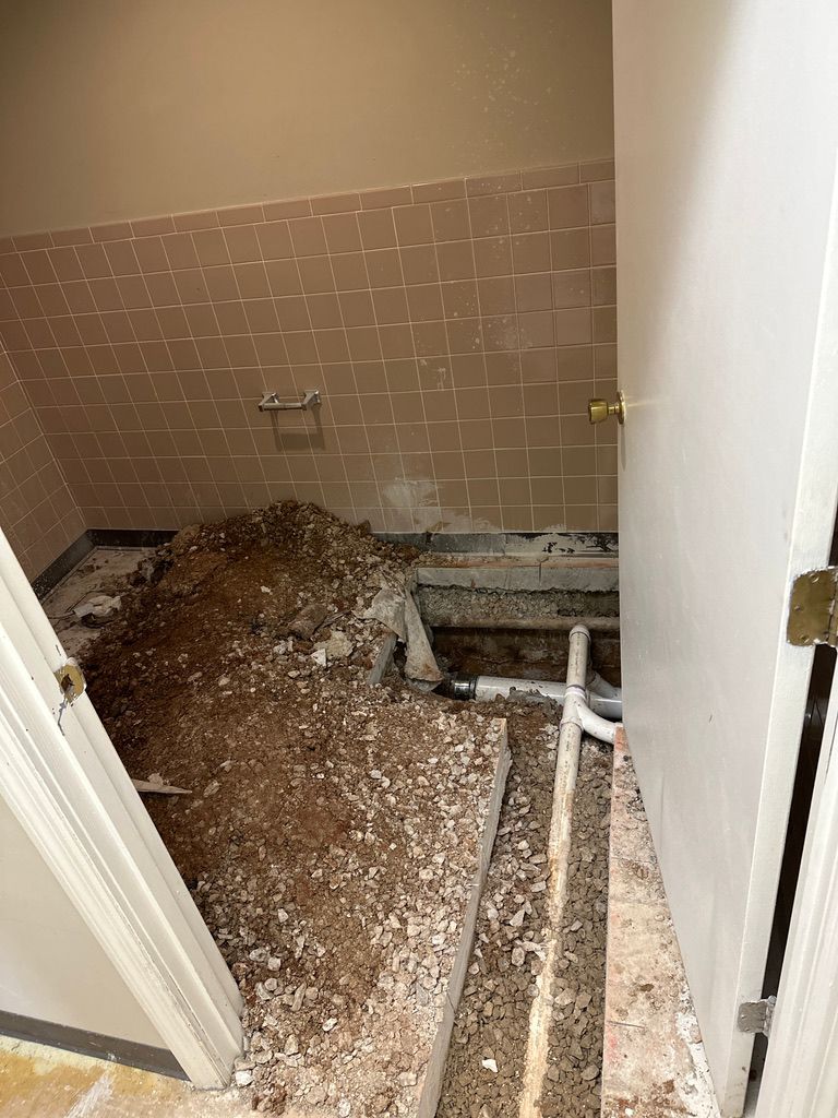 Bathroom floor torn up, exposing pipes and debris beside a tiled wall.