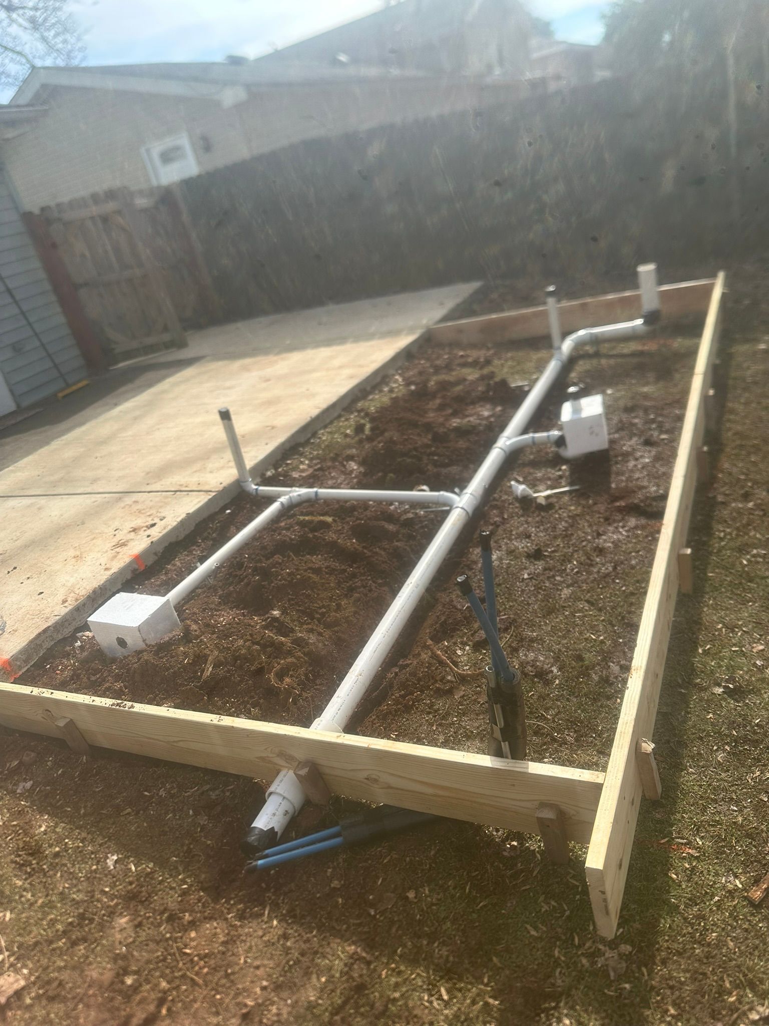 Raised garden bed frame with PVC pipes on soil in a backyard, viewed in sunlight