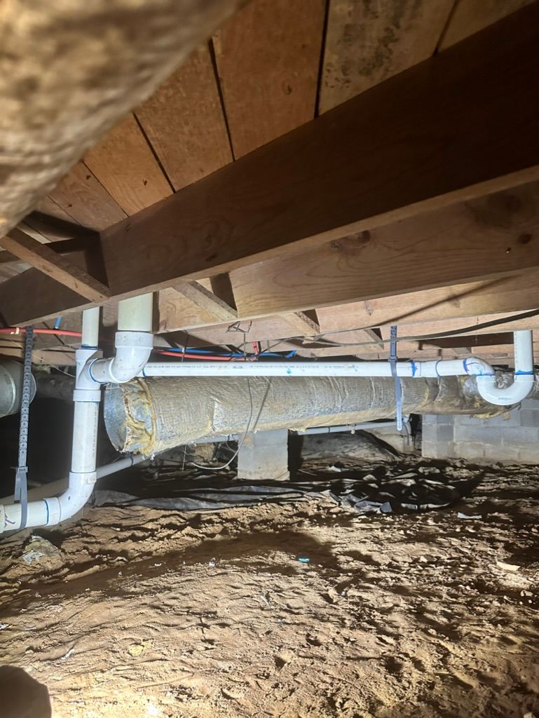 Crawl space under a house with exposed wooden beams, insulation, and white pipes over a dirt floor
