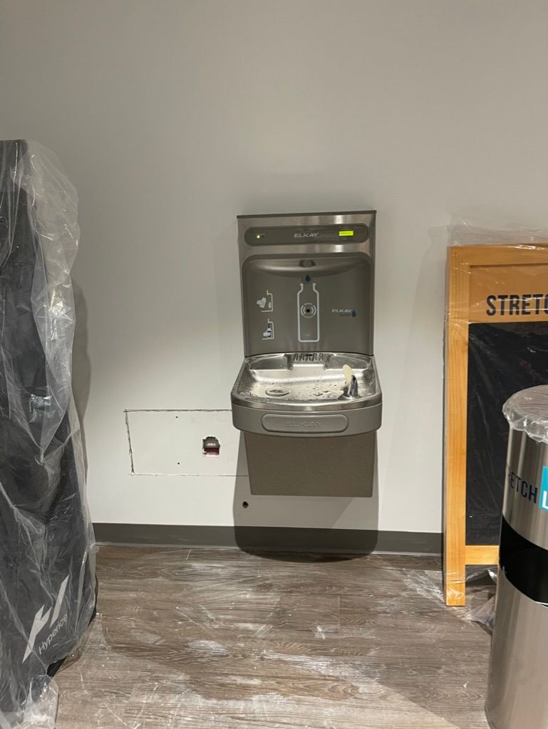 Public drinking fountain mounted on a gray wall in an indoor hallway, with wrapped items on both sides.