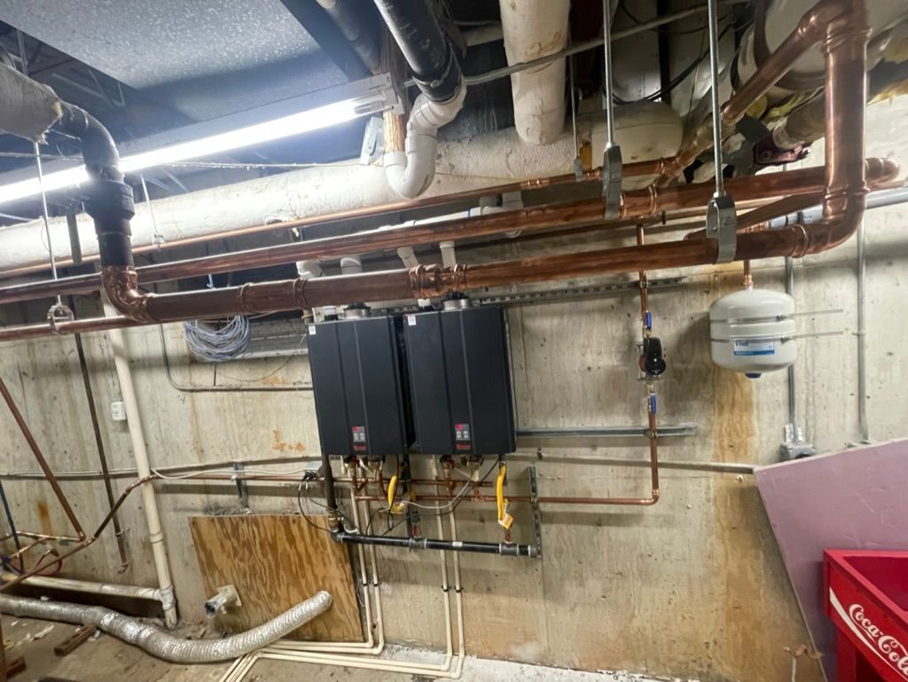 Basement utility room with exposed pipes, black electrical panels, and stained concrete walls